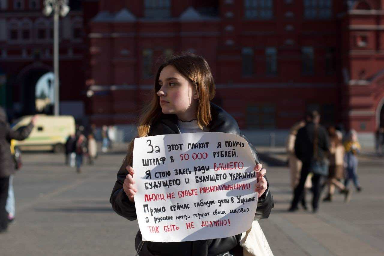 Una donna in piazza tiene in mano un cartello con una scritta in caratteri cirillici. Traduzione nella didascalia.